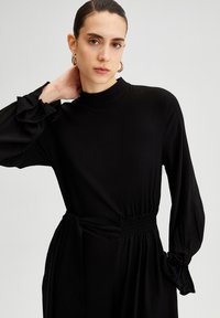 Rochie neagră cu mâneci lungi, gât înalt, talie elasticizată și mansete volan. Țesătură netedă și croială structurată, cu o siluetă elegantă.
