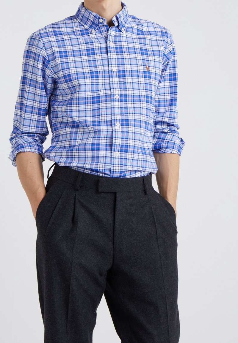 Blå och vit rutig skjorta med knäppning nedtill, kombinerad med mörkgrå byxor med veck och sidofickor.