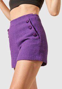 Shorts texturés violets avec une taille haute et des boutons latéraux. Présentent un motif tissé et une coupe ajustée, adaptés aux temps chauds.