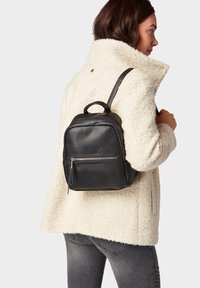 Petit sac à dos en cuir noir avec une surface texturée, poche zippée à l'avant et poignée sur le dessus. Porté par-dessus un manteau fluffy de couleur crème.
