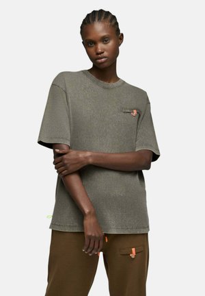 Jeune personne aux cheveux tressés portant un t-shirt gris ample et un pantalon marron avec des détails d'onglet orange, debout, les bras croisés.