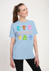 Světle modré tričko s barevnými pohádkov�ými medvědy a slovem "IKONICKÉ" napsaným tučnými, hravými písmeny přes přední část. Bavlněný materiál.