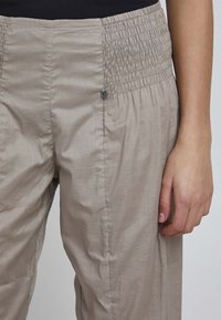 Beige lightweight trousers with a smocked waistband and subtle sheen. Features a decorative metal button and smooth texture.