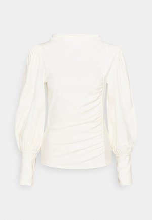 Biela blúzka s vysokým výstrihom, vypchatými dlhými rukávmi a bočnými nariaseniami. Hladká látka s jemnou textúrou.