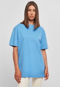 Žena se rovnými hnědými vlasy má na sobě oversized světle modré tričko s krátkým rukávem a krémové kalhoty, stojí na jednolitém šedém pozadí.