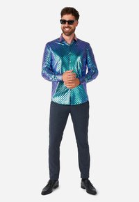 Camicia blu-verde brillante e iridescente con motivo a squame di pesce, design con bottoni, pantaloni neri e scarpe nere. Accessoriata con occhiali da sole.