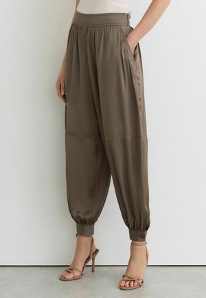 Femme portant un pantalon en satin vert olive ample, avec des chevilles retroussées et des poches, associé à des sandales à talons nude, debout contre un mur blanc.