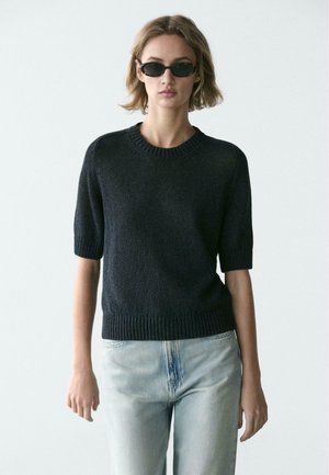 Kvinde iført sort kortærmet sweater, lyseblå jeans og små ovale sorte solbriller, stående foran en ensfarvet lys baggrund.