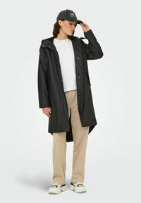 Frau, die eine schwarze "Los Angeles"-Kappe anpasst, trägt einen langen schwarzen Mantel, ein weißes Shirt, beige Hose und weiße Sneaker vor einem einfarbigen Hintergrund.