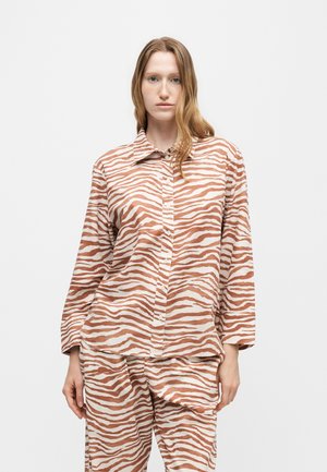 Kvinde med langt lysebrunt hår iført matchende løst zebra-stribet brun og hvid skjorte og bukser, stående mod en ensfarvet baggrund.