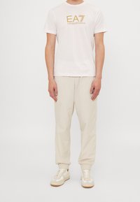 Vit t-shirt med guld "EA7 Emporio Armani"-logotyp, kombinerad med ljusbeiga elastiska byxor och vita sneakers med beige accenter.