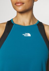 Una camiseta de deporte en color azul verdoso con detalles en negro, que presenta una superficie texturizada y el logo de "The North Face" en blanco en la zona del pecho.