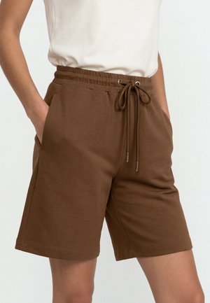 Brązowe elastyczne szorty z troczkiem w pasie, przednie kieszenie, noszone z białym bezrękawnikiem, ręce swobodnie w kieszeniach.