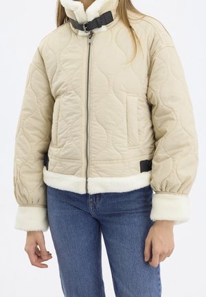 Veste matelassée beige avec doublure en shearling blanc, fermeture éclair à l’avant, col à boucle et poignets, portée avec un jean bleu sur une personne debout.