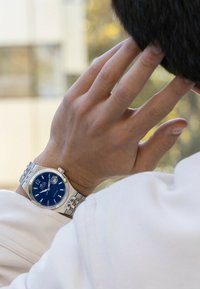 Srebrny zegarek ze stali nierdzewnej z niebieską tarczą, srebrnymi znacznikami i wyświetlaczem daty, noszony na nadgarstku, częściowo zasłonięty ręką.