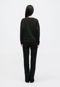 Černá mikina se zaobleným výstřihem a žebrovými manžetami, kombinovaná s černými kalhotami rovného střihu. Celkový design je minimalistický a jednobarevný.