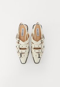 Hvite patentlær slingback-sko med spisse tær, tre justerbare stropper, sølvfarge detaljer og en sort detaljert såle.
