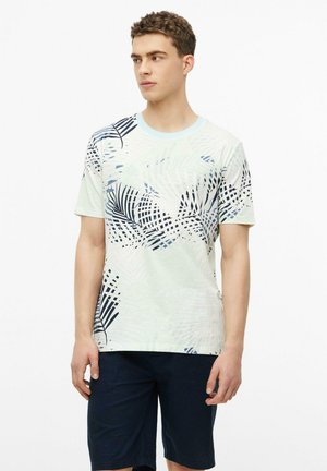 Jeune homme portant une chemise à manches courtes de couleur claire avec des motifs de feuilles de palmier bleues et un short bleu foncé, debout et regardant vers la gauche.