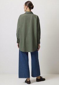 Grön långärmad skjorta med ryggpåsättning, krökt nederkant och knappmanschetter, i kombination med vida blå denimbyxor och bruna skor.