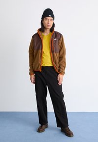 Chaqueta de forro polar marrón con detalles naranjas, camiseta amarilla, pantalones negros y botas oscuras. Modelo lleva un gorro de punto negro. Fondo sencillo.