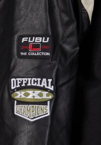 Sort læderjakke med FUBU og XXL broderede mærker, tekstureret overflade og kontrasterende cremefarvede detaljer.