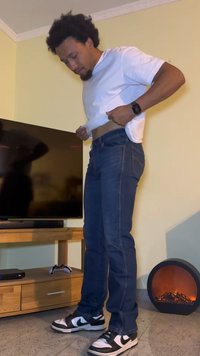 Jean en denim bleu avec une coupe droite, associé à un t-shirt blanc et des baskets noir et blanc. La pièce dispose d'une télévision et d'un radiateur décoratif.