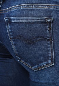 Jeans de mezclilla azul oscuro con un bolsillo trasero que presenta un patrón de costura curvado y una pequeña etiqueta con un logo. La tela tiene una textura suave.