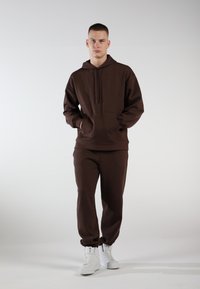 Brauner Kapuzenpullover und passende Jogginghose aus weichem Stoff. Mit Kängurutasche, Kordelzugkapuze und elastischen Bündchen. Ein Paar weiße Sneaker.