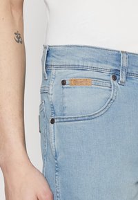 Világoskék Wrangler farmernadrág, fehér inggel, amelyen látható az első zsebek részlete és egy kar, amelyen fekete Om tetoválás található.