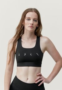 Svart sportbh med rundad halsringning, med en vit abstrakt logotyp över framsidan. Slät textur och åtsittande design.