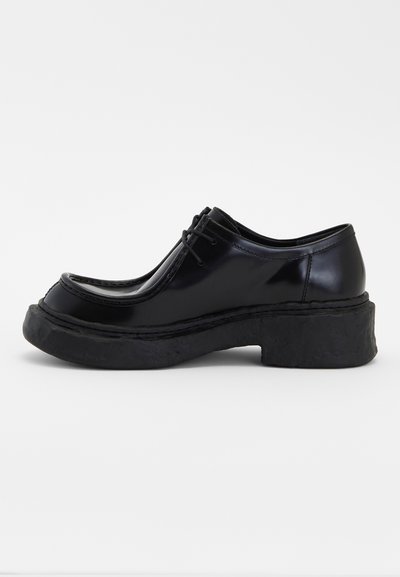 Zapatos de charol negro con punta redondeada, diseño de cordones y una suela de plataforma negra texturizada y gruesa. Costuras en el cuerpo del zapato.
