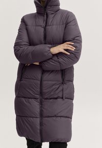 Osoba nosząca długą, ciemnofioletową puchową kurtkę z wysokim kołnierzem, zapinaną na zamek z przodu, z rękami skrzyżowanymi na piersi.