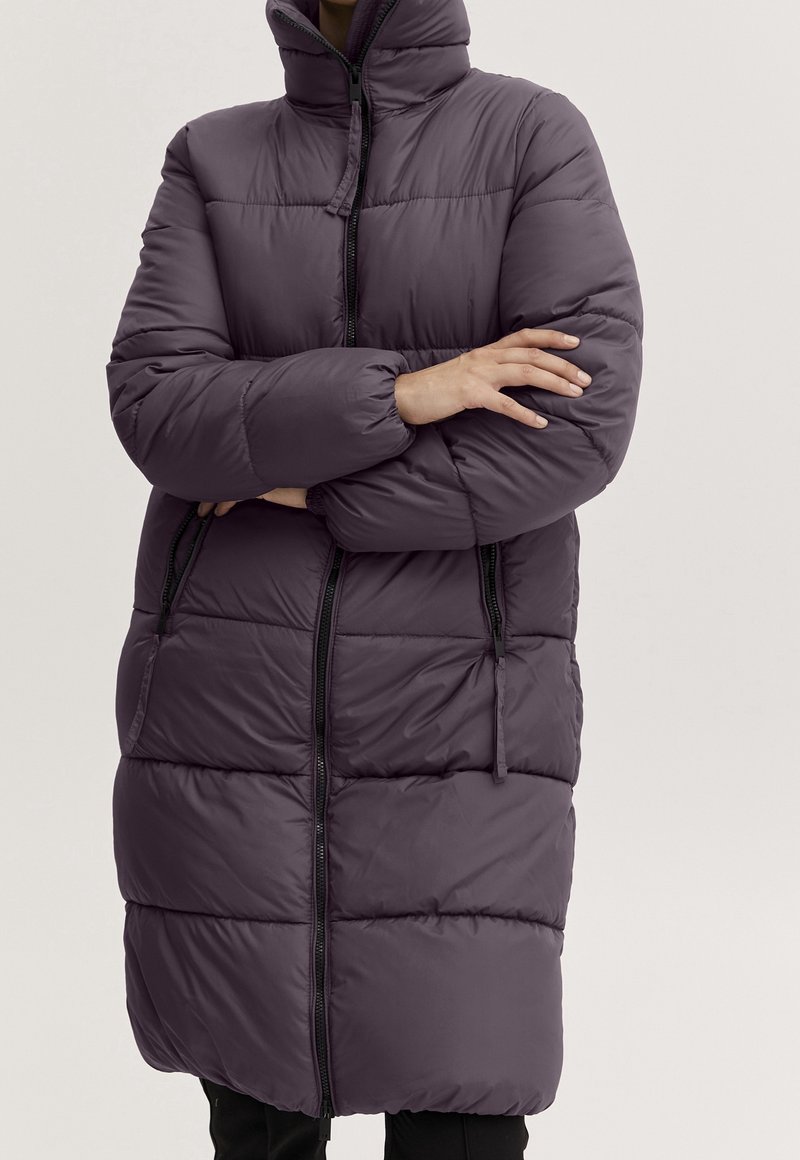 Osoba nosząca długą, ciemnofioletową puchową kurtkę z wysokim kołnierzem, zapinaną na zamek z przodu, z rękami skrzyżowanymi na piersi.