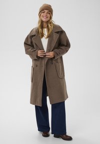 Palton din amestec de lână de culoare maro, cu reveruri mari, nasturi dublu închisi și buzunare laterale, asortat cu pantaloni largi de culoare bleumarin și o căciulă tricotată.