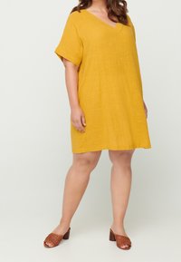 Gul kortärmad klänning med V-ringning och avslappnad passform, tillverkad av texturerad tyg. Kombinerad med bruna flätade klacksandaler.