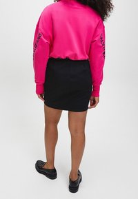 Růžová mikina se černým logem na rukávech, spárovaná s černou denimovou mini sukní a černými plochými botami. Oblečení má neformální střih.