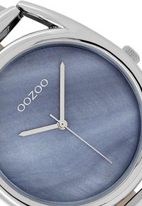 Montre ronde en argent avec un boîtier en métal poli, un cadran dégradé bleu, des aiguilles argentées et le logo 'oozoo'. Présente un design minimaliste.