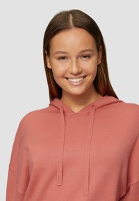 Jasnoróżowa bluza z kapturem, z dekoltem w kształcie V i sznurkami, wykonana z prążkowanego materiału. Posiada luźny krój i długie rękawy.