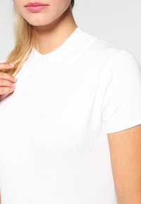 Närbild av en person som bär en vit ribbad kortärmad topp och rör vid sitt blonda hår vid axeln.