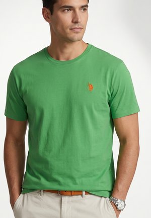 Uomo con t-shirt verde a maniche corte con piccolo logo arancione sul petto, pantaloni beige, cintura marrone e orologio da polso argentato, mani in tasca.