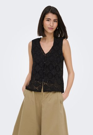 Femeie cu păr închis la culoare până la umeri, purtând un top negru din dantelă fără mâneci și pantaloni bej largi, cu mâinile în buzunare.