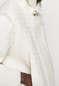 Biała kurtka o fakturowanej powierzchni z zapięciem na zamek, z kapturem i związanym detalem. Materiał ma lekko wypukły wzór i postrzępione krawędzie.