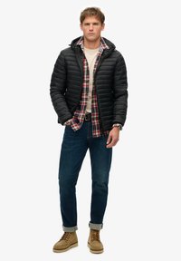 Sort pufferjakke med hætte, grå crewneck skjorte, rød og ternet flannel, blå jeans og brune støvler med snørebånd. Casual lagdeling.