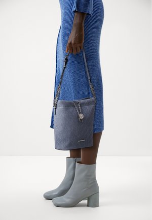 Jeanstasche im Beuteldesign mit strukturierter Oberfläche, ausgestattet mit einem Kordelzugverschluss, einem Kettenhenkel und einem Metallakzent. Kombiniert mit einem blauen Kleid und grauen Stiefeln.