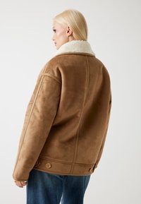 Giacca in faux suede marrone con un morbido colletto in shearling bianco, caratterizzata da una vestibilità ampia e grandi bottoni laterali. La vista posteriore mostra i dettagli delle cuciture.