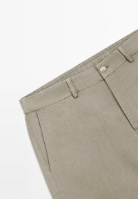 Beżowe spodnie z przednimi kieszeniami, szlufkami na pasek, zapięciem na guzik oraz elastycznym paskiem w talii na białym tle.