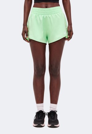 Personne athlétique portant un short de course vert clair, des baskets noires, des chaussettes blanches et un top court noir, debout devant un fond uni.