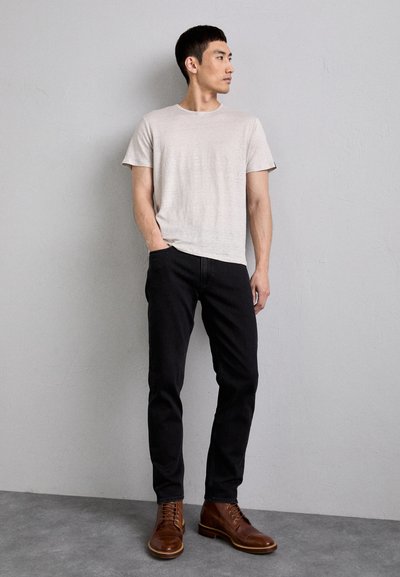 Camiseta de manga corta gris claro, jeans oscuros ajustados y botas de cuero marrón con cordones. El modelo está de pie frente a una pared texturizada.