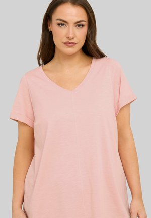 Frau mit langen braunen Haaren, die ein hellrosa kurzärmeliges V-Ausschnitt-Shirt vor einem schlichten grauen Hintergrund trägt.