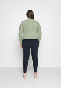 Pull ribbed vert à manches longues porté sur un jean skinny bleu foncé. Des chaussures de couleur claire avec des brides à la cheville complètent la tenue.